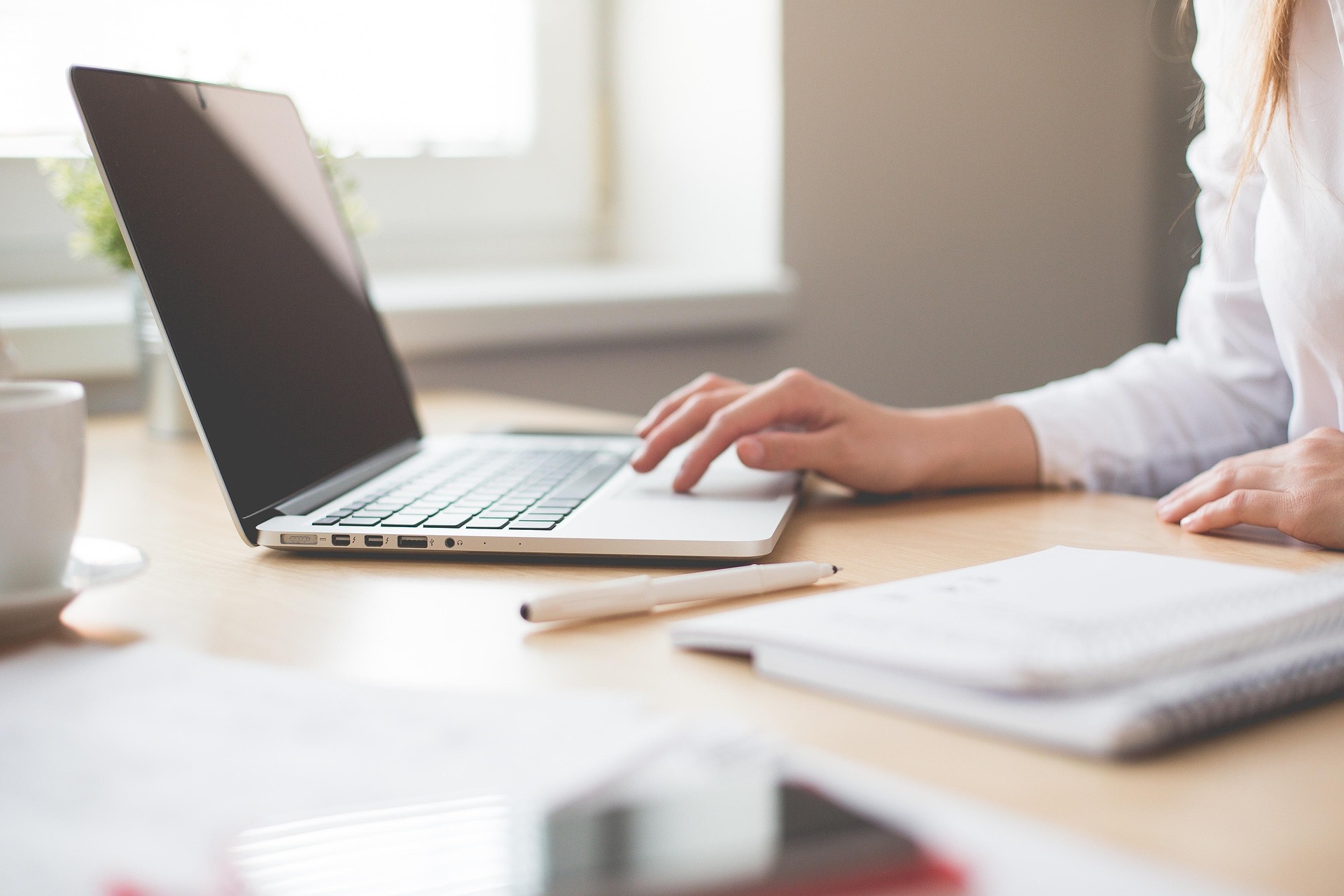 Person arbeitet am Laptop auf Holzschreibtisch mit Notizbuch und Kaffeetasse
