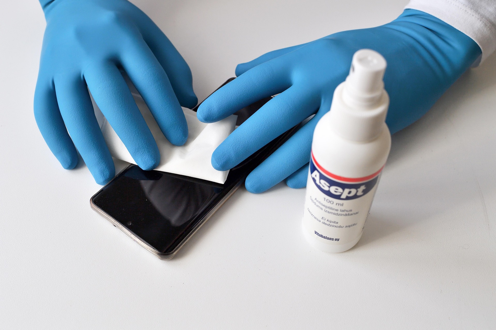 Hände mit blauen Einweghandschuhen desinfizieren Smartphone mit Tuch und Desinfektionsspray